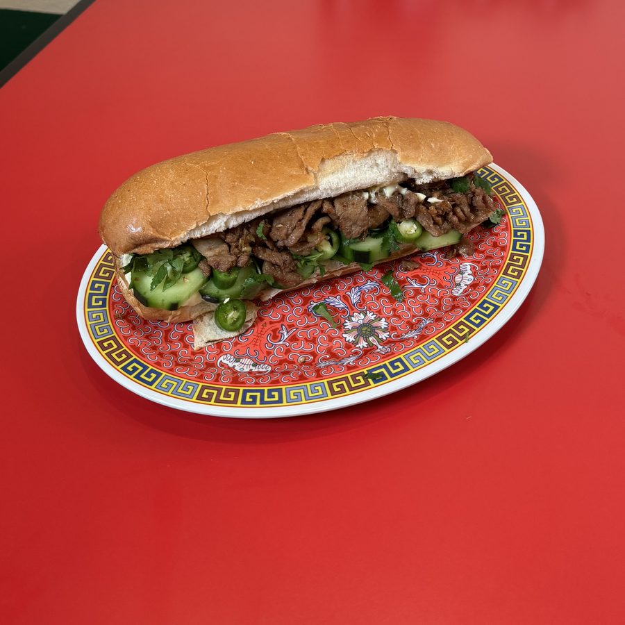 A banh mi on a french roll filled with sliced beef, cucumber and jalapeno, on a patterned oval plate on a red table.
