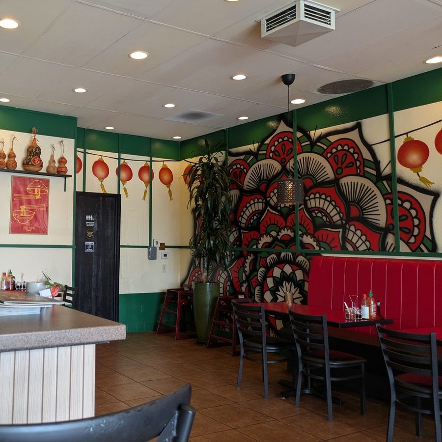 The dining room seen past the service counter, with green and cream wall panels, a mandala mural, hanging red lanterns and a red banquette behind the tables.