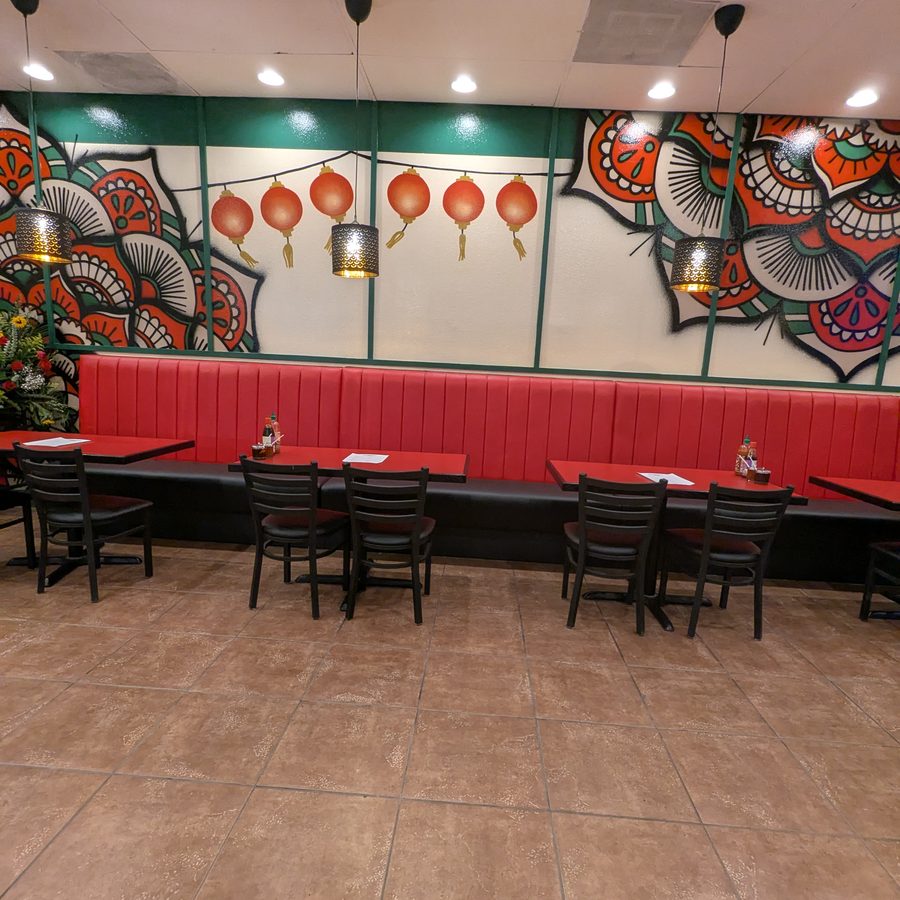 A long red banquette against a wall painted with red and green mandala murals, hung with red paper lanterns, with black chairs at red tables on a tile floor.
