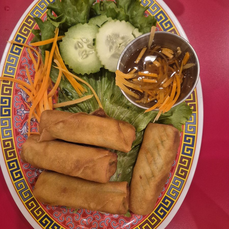 An oval plate with a red and gold rim holding fried egg rolls and tofu on lettuce, with cucumber slices, shredded carrot and a metal cup of dipping sauce.