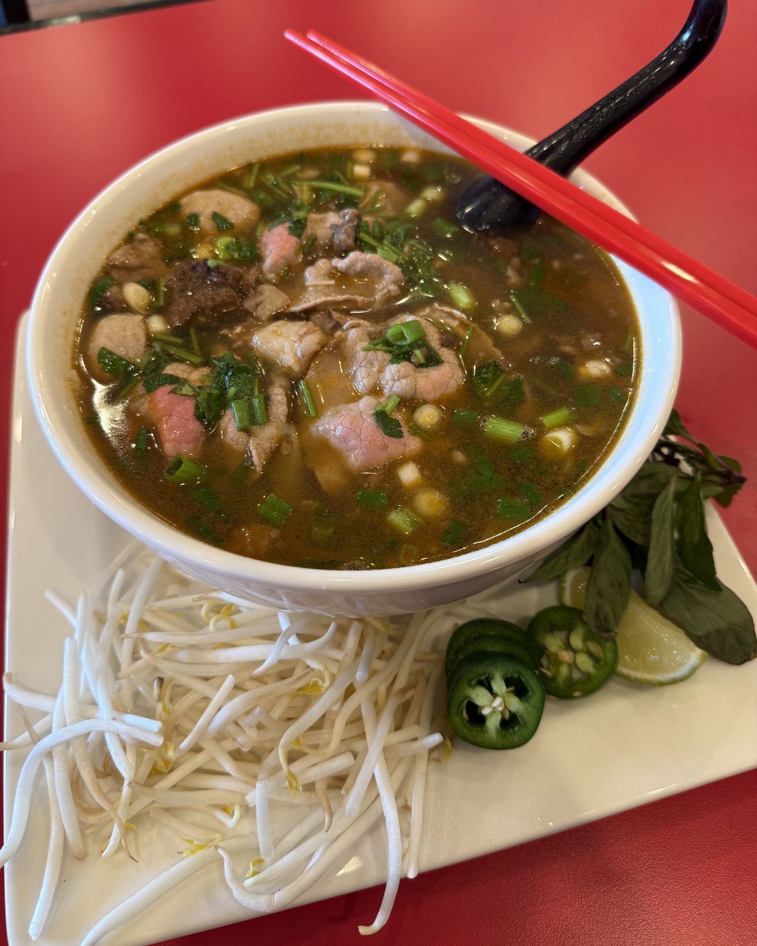 A white bowl of beef pho on a red table, filled with sliced beef, brisket, meatballs, scallions and cilantro, with red chopsticks and a black spoon across the rim and a plate of bean sprouts, basil, lime and sliced jalapeno beside it.