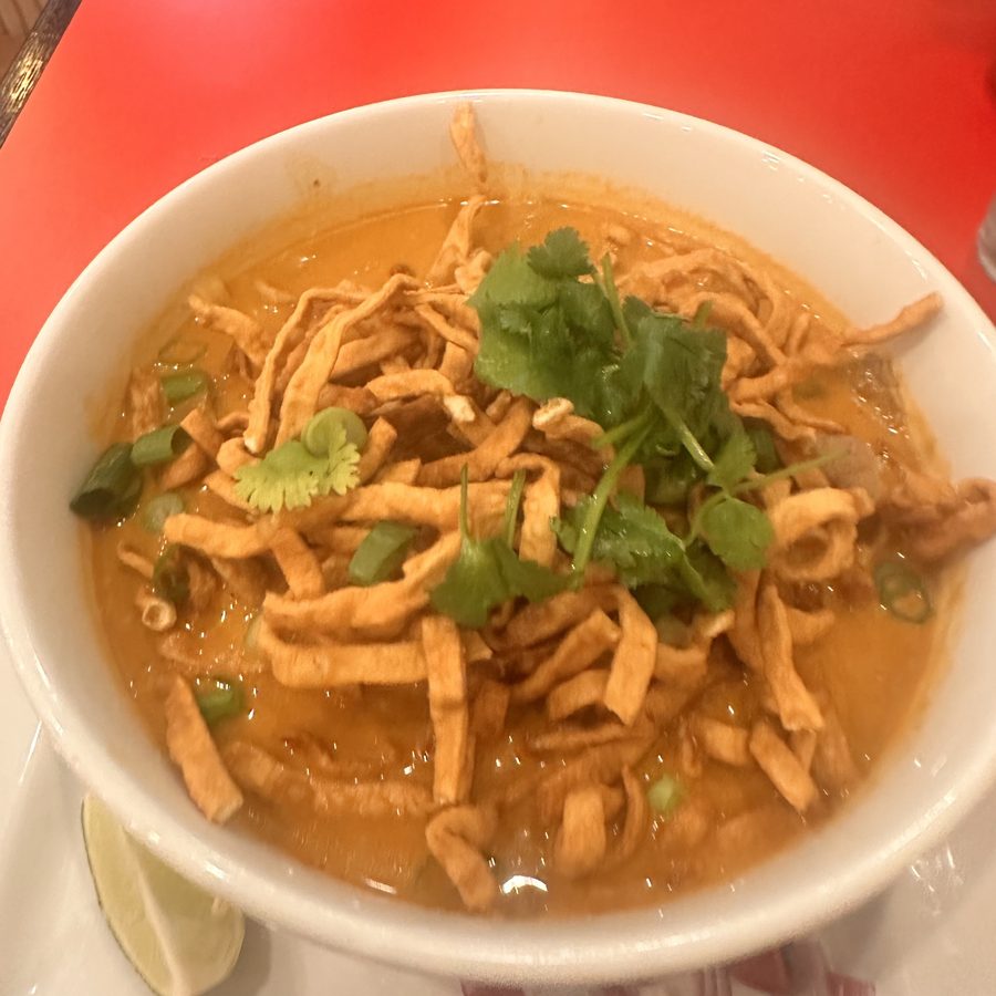 A white bowl of khao soi in orange coconut broth under a nest of crisp fried egg noodles, topped with cilantro and green onion.