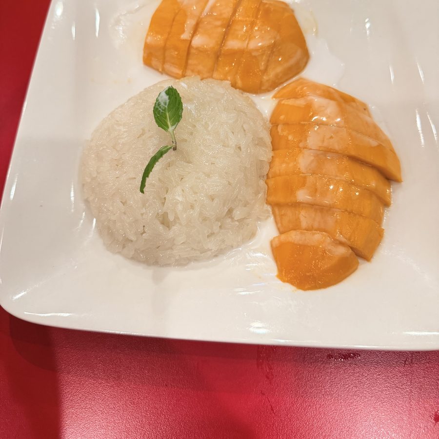 A white square plate holding a mound of sticky rice topped with a mint leaf, with sliced mango beside it.
